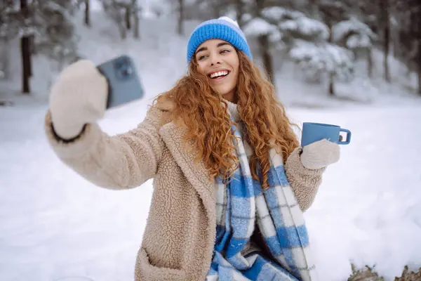 Portrait of a beautiful woman in warm clothes takes a selfie with a hot drink from a thermos in her hands. Happy female traveler with phone enjoying winter day outdoors.