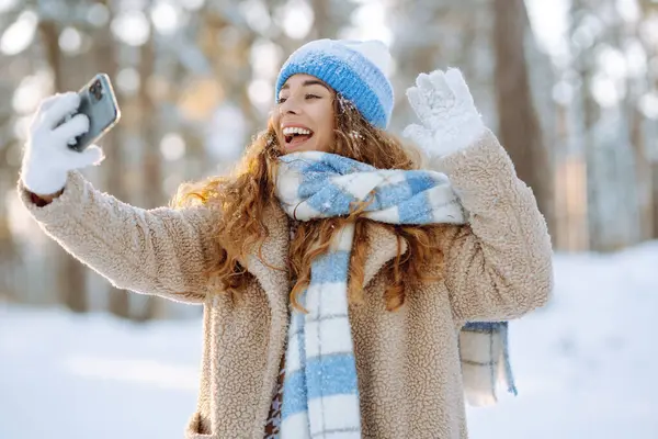 Karlı bir ormanda telefonu olan parlak şapkalı ve atkılı genç bir kadın. Dışarıdaki telefonundan mutlu kadın turist blogları. Seyahat, tatil konsepti.