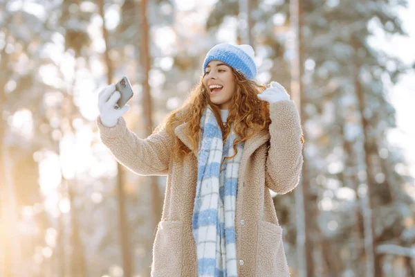 Karlı bir ormanda telefonu olan parlak şapkalı ve atkılı genç bir kadın. Dışarıdaki telefonundan mutlu kadın turist blogları. Seyahat, tatil konsepti.