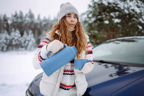 Attractive woman in a hat and gloves drinks a drink from a thermos near a car on a snowy road. Snowy forest. Car travel concept, freedom.