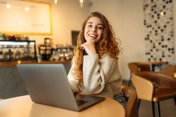 Young woman works with a laptop while sitting in a cafe. Freelancer works online in a modern coffee shop. Concept of technology, freelancing. Lifestyle.