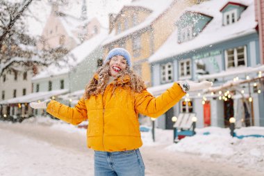 Parlak ceketli gülümseyen kadın karlı bir caddede karla eğleniyor. Genç bir kadın açık havada karlı havanın tadını çıkarıyor. Rahatlama kavramı, eğlence. Yaşam biçimi.