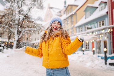 Parlak ceketli gülümseyen kadın karlı bir caddede karla eğleniyor. Genç bir kadın açık havada karlı havanın tadını çıkarıyor. Rahatlama kavramı, eğlence. Yaşam biçimi.