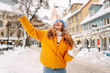 Parlak ceketli gülümseyen kadın karlı bir caddede karla eğleniyor. Genç bir kadın açık havada karlı havanın tadını çıkarıyor. Rahatlama kavramı, eğlence. Yaşam biçimi.