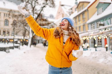 Parlak ceketli gülümseyen kadın karlı bir caddede karla eğleniyor. Genç bir kadın açık havada karlı havanın tadını çıkarıyor. Rahatlama kavramı, eğlence. Yaşam biçimi.