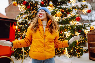 Parlak sarı ceketli ve kırmızı dudaklı mutlu kadın Noel pazarında yürüyor. Kış tatilinin tadını çıkaran kıvırcık saçlı turist kadın. Her yer aydınlık. Tatil kavramı, eğlence.