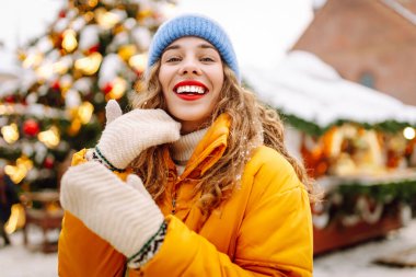 Parlak sarı ceketli ve kırmızı dudaklı mutlu kadın Noel pazarında yürüyor. Kış tatilinin tadını çıkaran kıvırcık saçlı turist kadın. Her yer aydınlık. Tatil kavramı, eğlence.