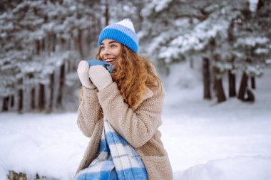 Karlı bir ormanda termosla gülen bir kadın doğanın tadını çıkarıyor. Dışarıda sıcak bir şeyler içen genç bir kadın. Macera konsepti. Yaşam biçimi.