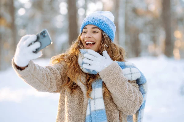 Sıcak kıyafetler içinde, elinde telefonla kışın karlı geçen bir günde eğlenen mutlu bir kadın. Genç bir kadın karlı bir parkta telefonuyla selfie çekiyor. Eğlenceli bir konsept. Etkin yaşam biçimi.