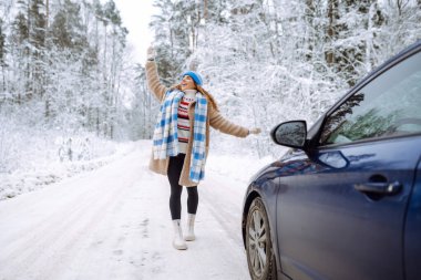 Karlı bir ormanda bir arabanın yanında güzel bir genç kadın. İlk kar, harika bir ruh hali. Bir gezgin doğada bir arabanın yanında poz verir. Araba yolculuğu, tatil kavramı.