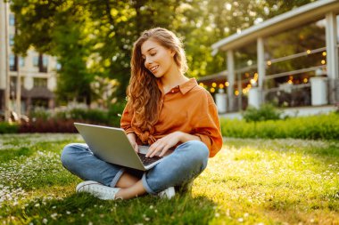 Güneşli bir parkta, yeşil çimlerde dizüstü bilgisayarla mutlu bir kadının portresi. Dizüstü bilgisayarda çalışan genç serbest çalışan kadın, güneşli havanın tadını çıkarıyor. Serbest konsept, teknoloji.