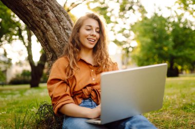 Güneşli bir parkta, yeşil çimlerde dizüstü bilgisayarla mutlu bir kadının portresi. Dizüstü bilgisayarda çalışan genç serbest çalışan kadın, güneşli havanın tadını çıkarıyor. Serbest konsept, teknoloji.