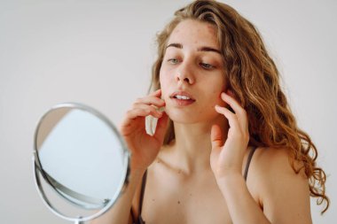 Close-up of a cute woman with problem skin looking in the mirror. A curly woman with rashes on her face is worried about the health of her skin. Dermatology concept, allergy.