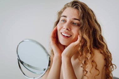 Close-up of a cute woman with problem skin looking in the mirror. A curly woman with rashes on her face is worried about the health of her skin. Dermatology concept, allergy.