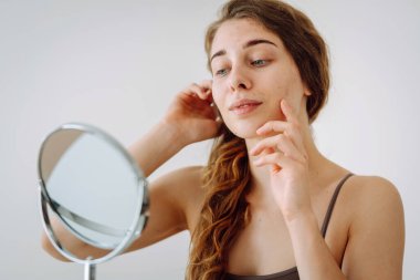 Close-up of a cute woman with problem skin looking in the mirror. A curly woman with rashes on her face is worried about the health of her skin. Dermatology concept, allergy.
