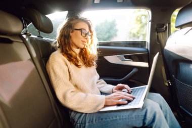İş ulaşımı. Dizüstü bilgisayarı olan kıvırcık saçlı bir kadın arabanın arka koltuğunda çalışıyor. Ulaşım ve teknoloji kavramı.