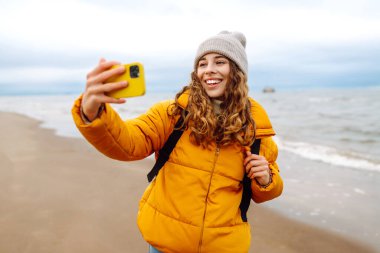 Deniz kıyısında telefonu olan mutlu bir kadının portresi. O parlak sarı bir ceket giyiyor. Genç bir kadın selfie çekiyor ve dışarıdaki manzaranın tadını çıkarıyor. Macera konsepti, tatil.