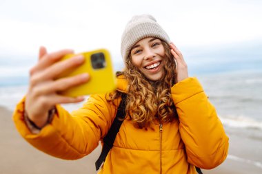 Deniz kıyısında telefonu olan mutlu bir kadının portresi. O parlak sarı bir ceket giyiyor. Genç bir kadın selfie çekiyor ve dışarıdaki manzaranın tadını çıkarıyor. Macera konsepti, tatil.