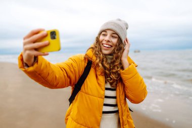 Deniz kıyısında telefonu olan mutlu bir kadının portresi. O parlak sarı bir ceket giyiyor. Genç bir kadın selfie çekiyor ve dışarıdaki manzaranın tadını çıkarıyor. Macera konsepti, tatil.
