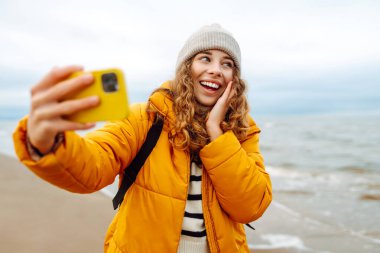 Deniz kıyısında telefonu olan mutlu bir kadının portresi. O parlak sarı bir ceket giyiyor. Genç bir kadın selfie çekiyor ve dışarıdaki manzaranın tadını çıkarıyor. Macera konsepti, tatil.
