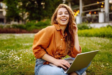 Hoş bir kadın dışarıda dizüstü bilgisayarla vakit geçiriyor. Neşeli kadın parktaki çimlerde internetten çalışıyor. Bağımsız çalışma kavramı, yürüme.