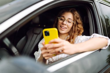 Bir kadın sürücünün portresi araba sürerken akıllı telefon kullanır. Arabadaki genç bir kadın yön bulmak için cep telefonu kullanıyor. Hareket konsepti, modern teknoloji.