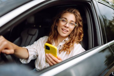 Bir kadın sürücünün portresi araba sürerken akıllı telefon kullanır. Arabadaki genç bir kadın yön bulmak için cep telefonu kullanıyor. Hareket konsepti, modern teknoloji.