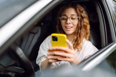 Bir kadın sürücünün portresi araba sürerken akıllı telefon kullanır. Arabadaki genç bir kadın yön bulmak için cep telefonu kullanıyor. Hareket konsepti, modern teknoloji.