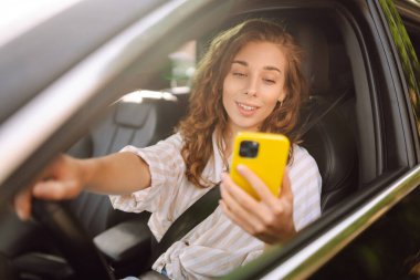 Bir kadın sürücünün portresi araba sürerken akıllı telefon kullanır. Arabadaki genç bir kadın yön bulmak için cep telefonu kullanıyor. Hareket konsepti, modern teknoloji.