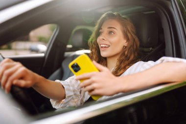 Bir kadın sürücünün portresi araba sürerken akıllı telefon kullanır. Arabadaki genç bir kadın yön bulmak için cep telefonu kullanıyor. Hareket konsepti, modern teknoloji.