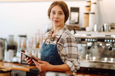 Modern bir kafede barın arkasında dijital tablet kullanan gülümseyen bir kafe sahibinin portresi. Genç bir barista tablet kullanarak sipariş alır. Küçük işletme kavramı.