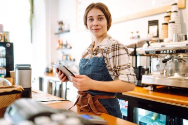 Modern bir kafede barın arkasında dijital tablet kullanan gülümseyen bir kafe sahibinin portresi. Genç bir barista tablet kullanarak sipariş alır. Küçük işletme kavramı.