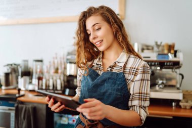 Modern bir kafede barın arkasında dijital tablet kullanan gülümseyen bir kafe sahibinin portresi. Genç bir barista tablet kullanarak sipariş alır. Küçük işletme kavramı.