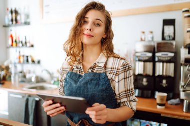 Modern bir kafede barın arkasında dijital tablet kullanan gülümseyen bir kafe sahibinin portresi. Genç bir barista tablet kullanarak sipariş alır. Küçük işletme kavramı.