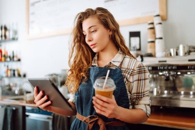 Modern bir kafede barın arkasında dijital tablet kullanan gülümseyen bir kafe sahibinin portresi. Genç bir barista tablet kullanarak sipariş alır. Küçük işletme kavramı.