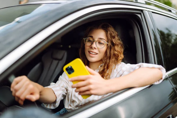 Bir kadın sürücünün portresi araba sürerken akıllı telefon kullanır. Arabadaki genç bir kadın yön bulmak için cep telefonu kullanıyor. Hareket konsepti, modern teknoloji.