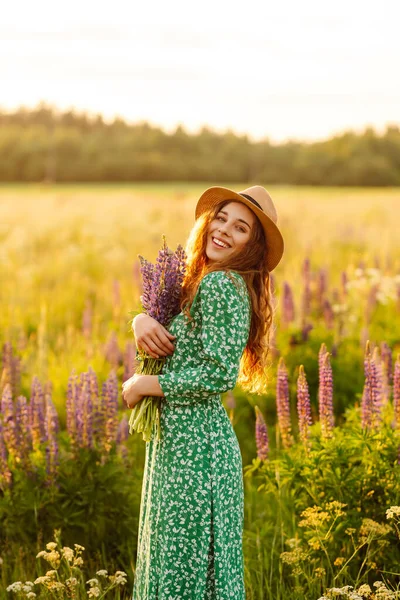 A young woman in a stylish green dress and hat enjoys the sunset in a field with lupins. A beautiful woman with a bouquet of lupins, collects flowers, inhales fresh floral air.