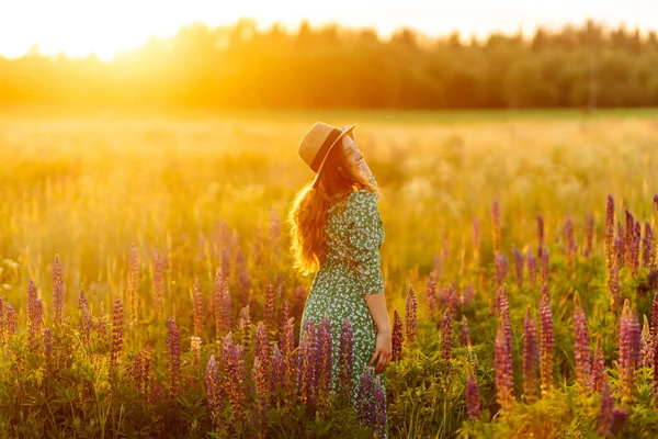A young woman in a stylish green dress and hat enjoys the sunset in a field with lupins. A beautiful woman with a bouquet of lupins, collects flowers, inhales fresh floral air.