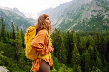 Sarı sırt çantalı mutlu kadın dağ manzarasının tadını çıkarıyor. Genç bir gezgin dağ patikaları boyunca seyahat eder. Macera, seyahat konsepti