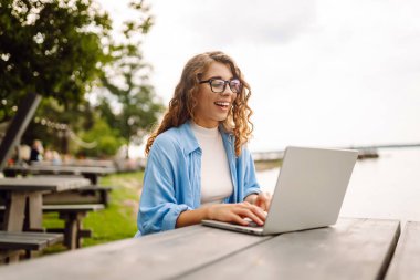 Şık giyinmiş, dışarıda bir masada oturmuş göl manzarası güzel olan bir dizüstü bilgisayarda çalışan genç bir kadın. Serbest çalışma konsepti. Çevrimiçi eğitim.