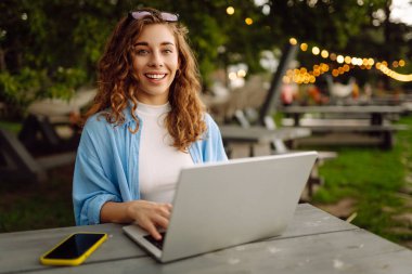 Şık giyinmiş, dışarıda bir masada oturmuş göl manzarası güzel olan bir dizüstü bilgisayarda çalışan genç bir kadın. Serbest çalışma konsepti. Çevrimiçi eğitim.