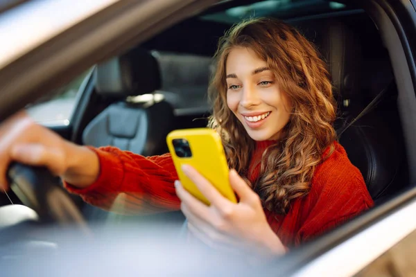 Portrait of a young woman sitting in a car in the driver\'s seat looking into a smartphone, paying for parking and navigating in the city. Concept of transport, mobility, carsharing.