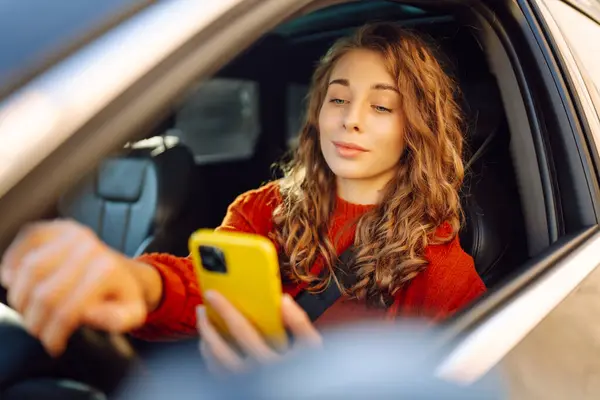 Portrait of a young woman sitting in a car in the driver\'s seat looking into a smartphone, paying for parking and navigating in the city. Concept of transport, mobility, carsharing.