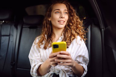 Genç bir kadın arabanın arka koltuğunda otururken akıllı telefon kullanır. İş, teknoloji, seyahat, online iletişim kavramı. Taksi..
