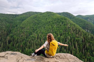 Sırt çantalı genç bir kadın dağın zirvesinde güzel bir dağa bakıyor. Yeşil tepeli, genç bir bayanla manzara. Yürüyüş. Doğa. Seyahat ve turizm.