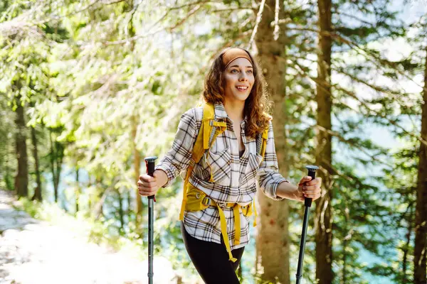 Young woman with hiking equipment walks along a hiking trail against the backdrop of mountain scenery. Summer vacation trip on weekend. Active lifestyle