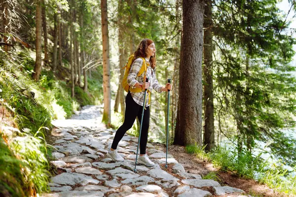 Young woman with hiking equipment walks along a hiking trail against the backdrop of mountain scenery. Summer vacation trip on weekend. Active lifestyle