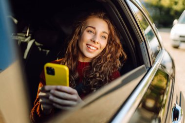 Genç bir kadın arabanın arka koltuğunda otururken akıllı telefon kullanır. İş, teknoloji, seyahat, çevrimiçi iletişim kavramı.