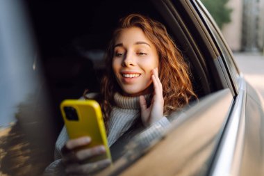 Genç bir kadın arabanın arka koltuğunda otururken akıllı telefon kullanır. İş, teknoloji, seyahat, çevrimiçi iletişim kavramı.