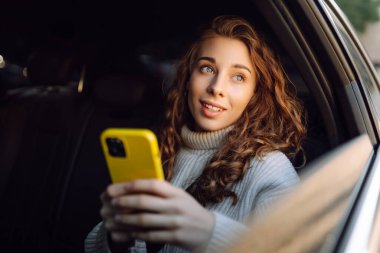 Genç bir kadın arabanın arka koltuğunda otururken akıllı telefon kullanır. İş, teknoloji, seyahat, çevrimiçi iletişim kavramı.